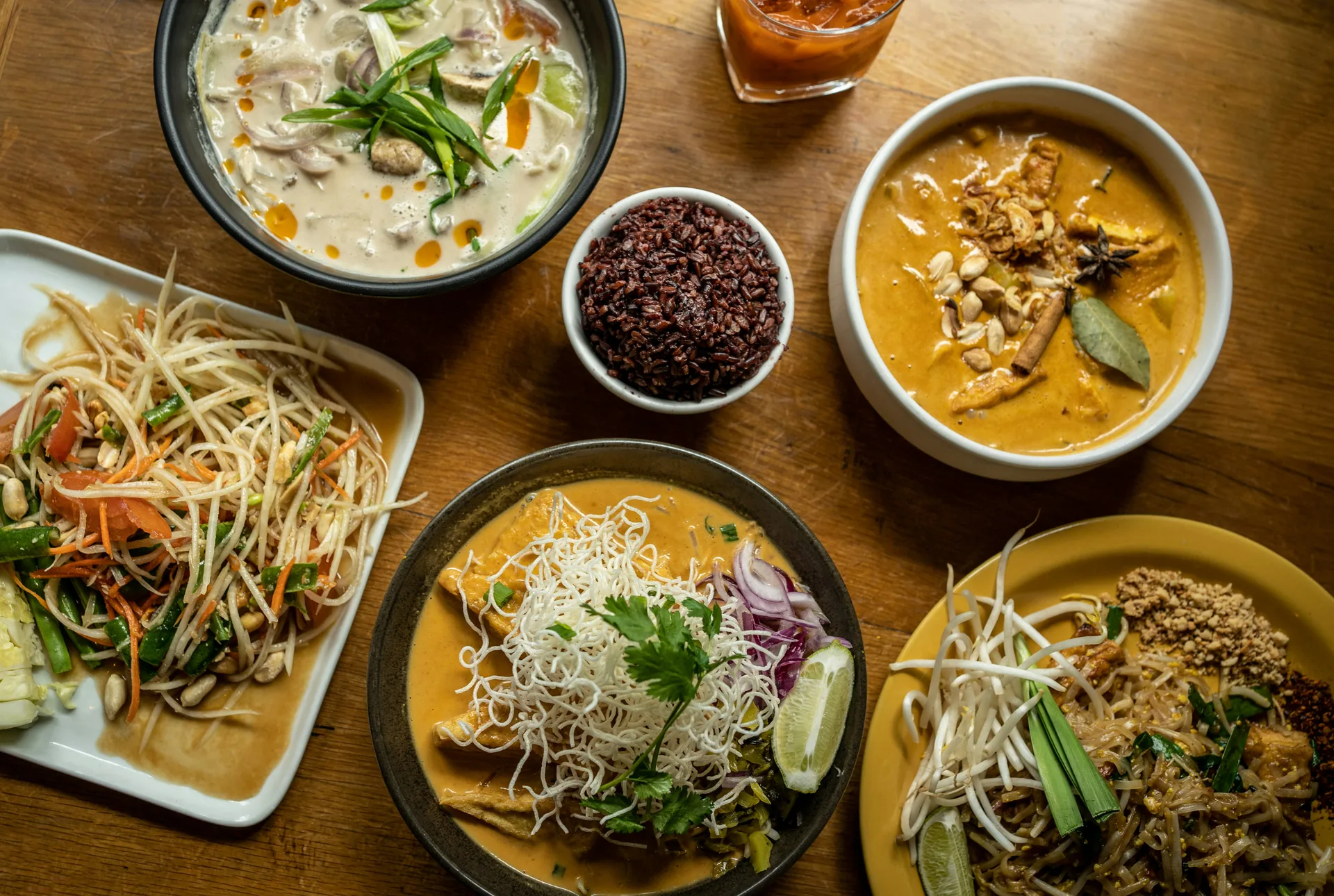 Spread of authentic Thai dishes on a wooden table, including green papaya salad, coconut chicken soup, yellow chicken curry, pad Thai, and a bowl of riceberry rice
